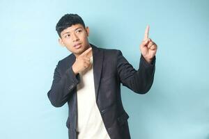 Portrait of young Asian business man in casual suit looking and pointing upward. Isolated image on blue background photo