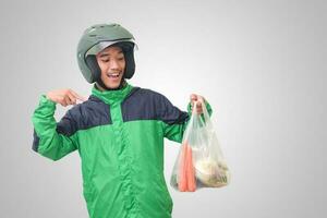 Portrait of Asian online taxi driver wearing green jacket and helmet delivering the vegetables from traditional market and pointing with finger. Isolated image on white background photo