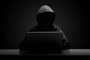 Mysterious man wearing black hoodie using laptop in the dark room. Hacker and cyber security concept. photo