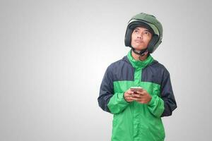 Portrait of confused Asian online taxi driver wearing green jacket and helmet thinking about an idea and looking up. Isolated image on white background photo