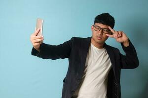 Portrait of young Asian business man in casual suit taking a selfie with confident and happy expression. Isolated image on blue background photo