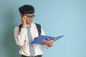 Indonesian senior high school student wearing white shirt uniform with gray tie writing on note book using pen and thinking about an idea. Isolated image on blue background photo