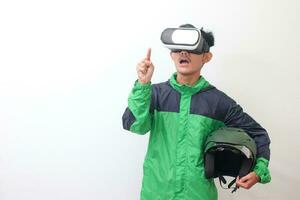 Portrait of Asian online taxi driver wearing green jacket and holding a helmet while using virtual reality or VR and pointing. Isolated image on white background photo