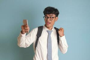 indonesio mayor alto colegio estudiante vistiendo blanco camisa uniforme con gris Corbata levantamiento su puño, celebrando éxito mientras participación un móvil teléfono. aislado imagen en azul antecedentes foto