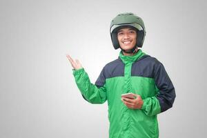 Portrait of Asian online taxi driver wearing green jacket and helmet holding mobile phone and pointing to empty space with finger. Isolated image on white background photo