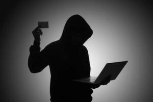 Mysterious man wearing black hoodie using laptop and holding credit card in the dark room. Hacker and cyber security concept. photo