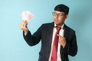 Portrait of young Asian business man in casual suit looking surprised and pointing at thousand rupiahs while holding it. Isolated image on blue background photo