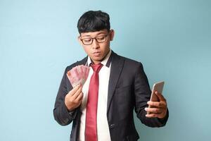 Portrait of young Asian business man in casual suit looking at camera while holding phone thousand rupiahs. Isolated image on blue background photo