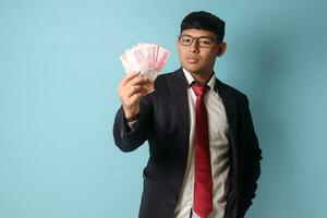 Portrait of young Asian business man in casual suit looking proud while holding thousand rupiahs. Isolated image on blue background photo