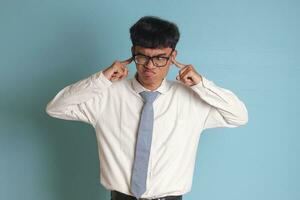 Indonesian senior high school student wearing white shirt uniform with gray tie covering his ears with his fingers, trying to avoid sounds or voices. Isolated image on blue background photo