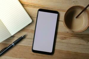 Blank screen mobile phone with empty notebook, pen, and a cup of coffee on wooden table during the day. Office work concept. Flat lay images photo