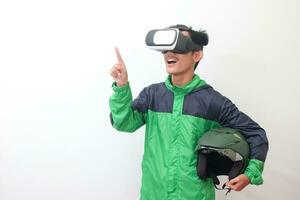 Portrait of Asian online taxi driver wearing green jacket and holding a helmet while using virtual reality or VR and pointing. Isolated image on white background photo