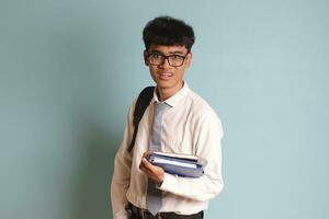 indonesio mayor alto colegio estudiante vistiendo blanco camisa uniforme con gris Corbata participación algunos libros, sonriente y mirando a cámara. aislado imagen en azul antecedentes foto