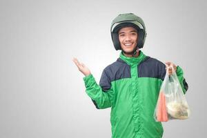 retrato de asiático en línea Taxi conductor vistiendo verde chaqueta y casco entregando el vegetales desde tradicional mercado y señalando con dedo. aislado imagen en blanco antecedentes foto