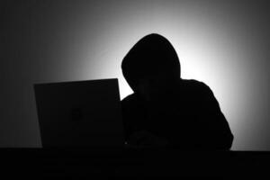 misterioso hombre vistiendo negro capucha utilizando ordenador portátil y participación crédito tarjeta en el oscuro habitación. hacker y ciber seguridad concepto. foto