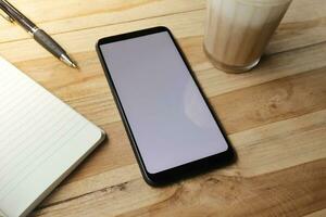 Blank screen mobile phone with empty notebook, pen, and a cup of coffee on wooden table during the day. Office work concept. Flat lay images photo