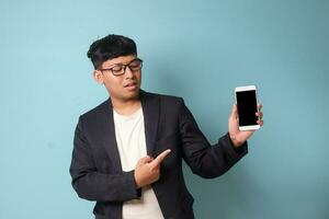 Portrait of young Asian business man in casual suit pointing at phone with confused expressions. Isolated image on blue background photo