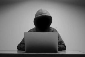 Mysterious man wearing black hoodie using laptop in the dark room. Hacker and cyber security concept. photo