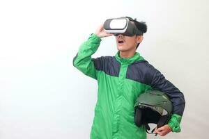 retrato de asiático en línea Taxi conductor vistiendo verde chaqueta y participación un casco mientras utilizando virtual realidad o vr y apuntando aislado imagen en blanco antecedentes foto