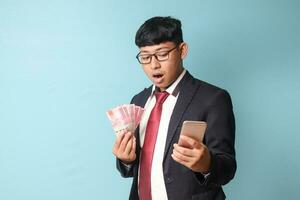 Portrait of young Asian business man in casual suit looking at smartphone surprised while holding thousand rupiahs. Isolated image on blue background photo