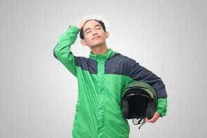 Portrait of tired Asian online taxi driver wearing green jacket straightening his hair and holding helmet, feeling exhausted. Isolated image on white background photo