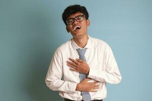 indonesio mayor alto colegio estudiante vistiendo blanco camisa uniforme con gris Corbata riendo ruidosamente mientras participación su pecho. divertido comedia concepto. aislado imagen en azul antecedentes foto