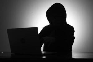 Mysterious man wearing black hoodie using laptop and holding credit card in the dark room. Hacker and cyber security concept. photo