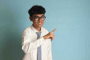 Indonesian senior high school student wearing white shirt uniform with gray tie showing product, pointing at something and smiling. Isolated image on blue background photo
