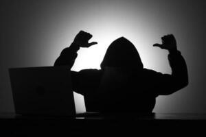 Mysterious man wearing black hoodie using laptop while mocking someone by making thumb down gesture in the dark room. Hacker and cyber security concept. photo