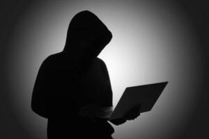 Mysterious man wearing black hoodie using laptop in the dark room. Hacker and cyber security concept. photo