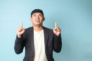 Portrait of young Asian business man in casual suit pointing and looking upward with happy expressions. Isolated image on blue background photo