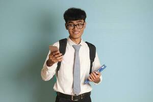 indonesio mayor alto colegio estudiante vistiendo blanco camisa uniforme con gris Corbata demostración conmocionado cara expresión mientras participación un móvil teléfono. aislado imagen en blanco antecedentes foto