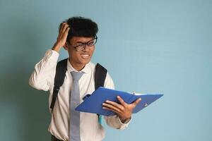 indonesio mayor alto colegio estudiante vistiendo blanco camisa uniforme con gris Corbata escritura en Nota libro utilizando bolígrafo y pensando acerca de un idea. aislado imagen en azul antecedentes foto