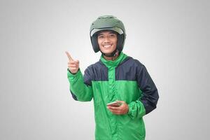 Portrait of Asian online taxi driver wearing green jacket and helmet holding mobile phone and pointing to empty space with finger. Isolated image on white background photo