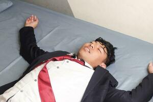 Portrait of young Asian business man in casual suit utterly defeated in bed with very exhausted expressions. Isolated image on blue background photo