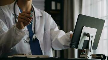 Serious female doctor using laptop and writing notes in medical journal sitting at desk. Young woman professional medic physician wearing white coat and stethoscope working on computer at workplace. photo