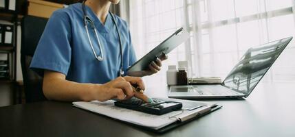 Serious female doctor using laptop and writing notes in medical journal sitting at desk. Young woman professional medic physician wearing white coat and stethoscope working on computer at workplace. photo