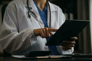 Serious female doctor using laptop and writing notes in medical journal sitting at desk. Young woman professional medic physician wearing white coat and stethoscope working on computer at workplace. photo