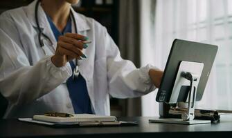 Serious female doctor using laptop and writing notes in medical journal sitting at desk. Young woman professional medic physician wearing white coat and stethoscope working on computer at workplace. photo