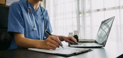 Serious female doctor using laptop and writing notes in medical journal sitting at desk. Young woman professional medic physician wearing white coat and stethoscope working on computer at workplace. photo