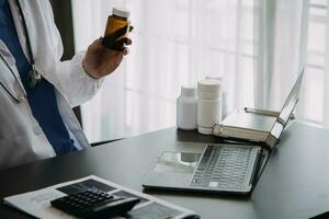 Serious female doctor using laptop and writing notes in medical journal sitting at desk. Young woman professional medic physician wearing white coat and stethoscope working on computer at workplace. photo