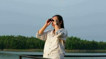 Young female explorer with binoculars exploring nature or watching birds outdoors. Young woman looking through binoculars at birds on the reservoir. Birdwatching video