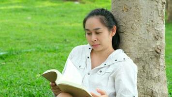 Relaxed young woman in a jacket reading a book in the summer park. Woman sitting on the grass and resting under the tree during vacation. Education concept video