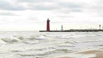 groot golven crashen op de strand met een rood vuurtoren in de in de buurt afstand gelegen Aan een pier in de oceaan. Aan een winderig bewolkt dag. video