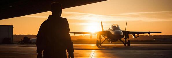 Sunset backlit view of military fighter jet pilot beside parked military airforce plane next to barracks or hangar as wide banner with copyspace area for world war conflicts. photo