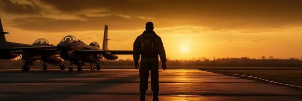 Sunset backlit view of military fighter jet pilot beside parked military airforce plane next to barracks or hangar as wide banner with copyspace area for world war conflicts. photo