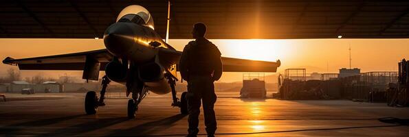 Sunset backlit view of military fighter jet pilot beside parked military airforce plane next to barracks or hangar as wide banner with copyspace area for world war conflicts. photo