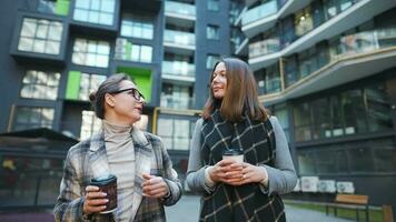 Two happy women walking with takeaway coffee and talking with interest among themselves in the business district. Slow motion video