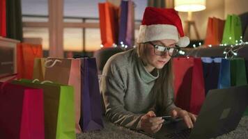 contento mujer con lentes vistiendo un Papa Noel claus sombrero es acostado en el alfombra y hace un en línea compra utilizando un crédito tarjeta video
