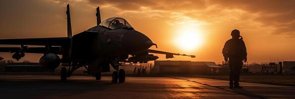 Sunset backlit view of military fighter jet pilot beside parked military airforce plane next to barracks or hangar as wide banner with copyspace area for world war conflicts. photo